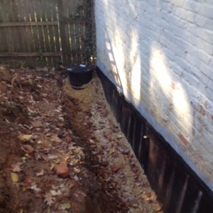 A narrow trench beside a white brick wall is filled with gravel, surrounded by fallen leaves. A dark cylindrical container sits at the trench's end.