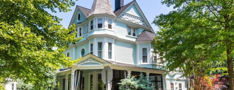 Victorian-style house with a turquoise exterior, ornate turret, and wraparound porch, surrounded by lush greenery under a clear blue sky.