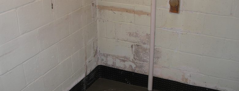 Basement corner with exposed concrete walls, a sump pump pit, and a white PVC pipe. The area appears utilitarian and slightly worn.